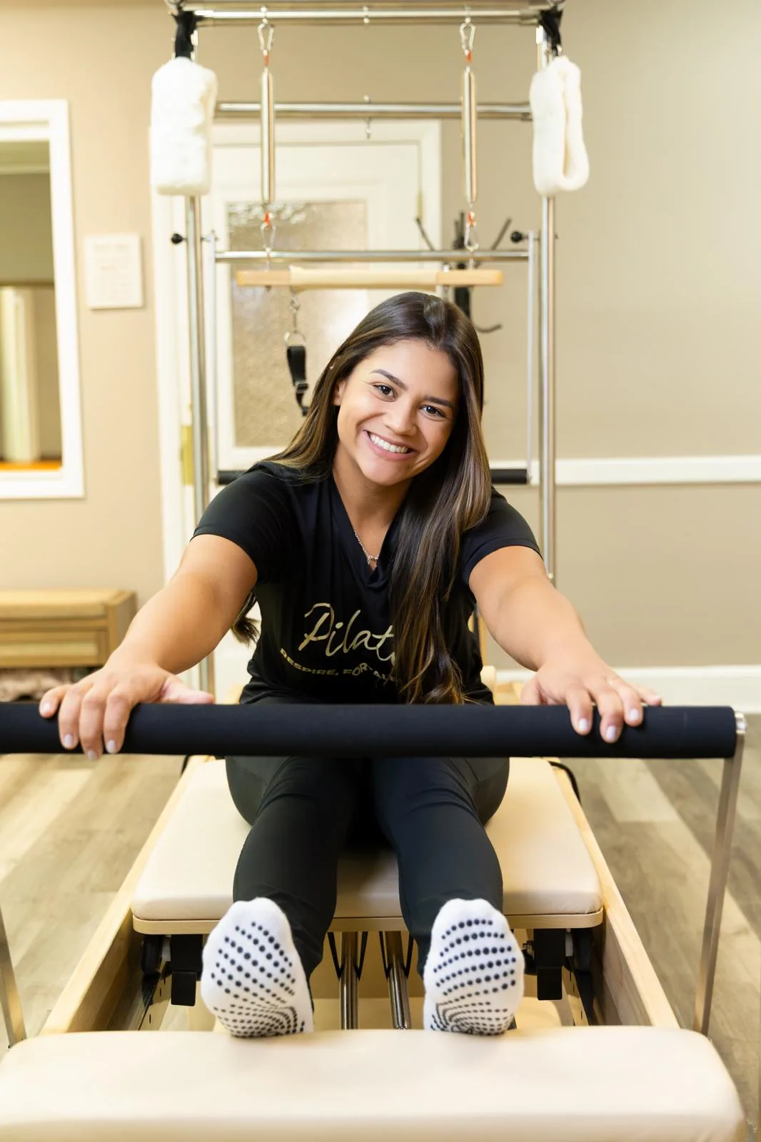 Instrutora com roupa de treino justa sentada no Reformer, segurando a barra de pés